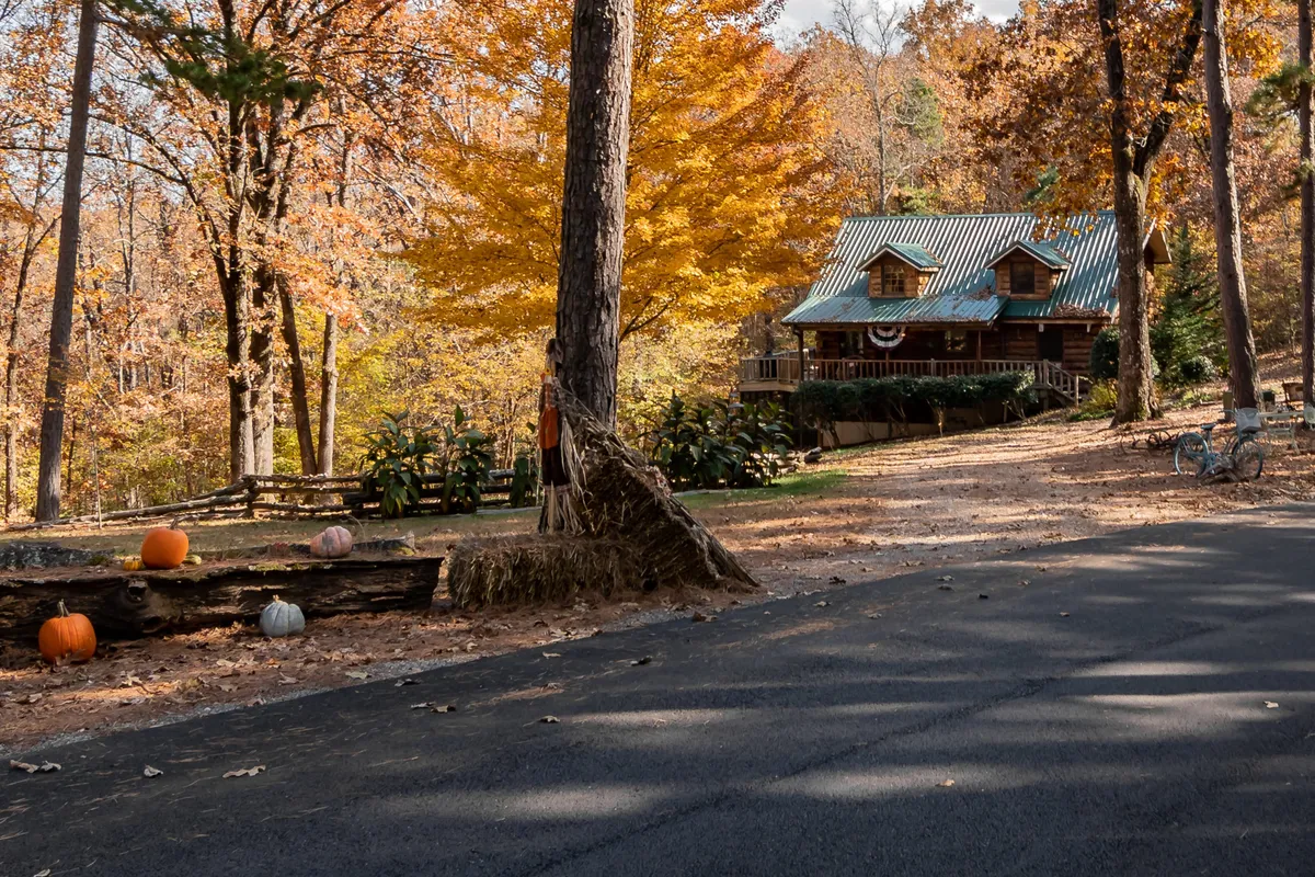 A log cabin surrounded by vibrant autumn foliage, decorated with pumpkins and a hay bale. The festive, seasonal touches evoke family gatherings and outdoor fun. The backdrop of colorful leaves and open space underscores the opportunity for nature walks and explorations during fall.