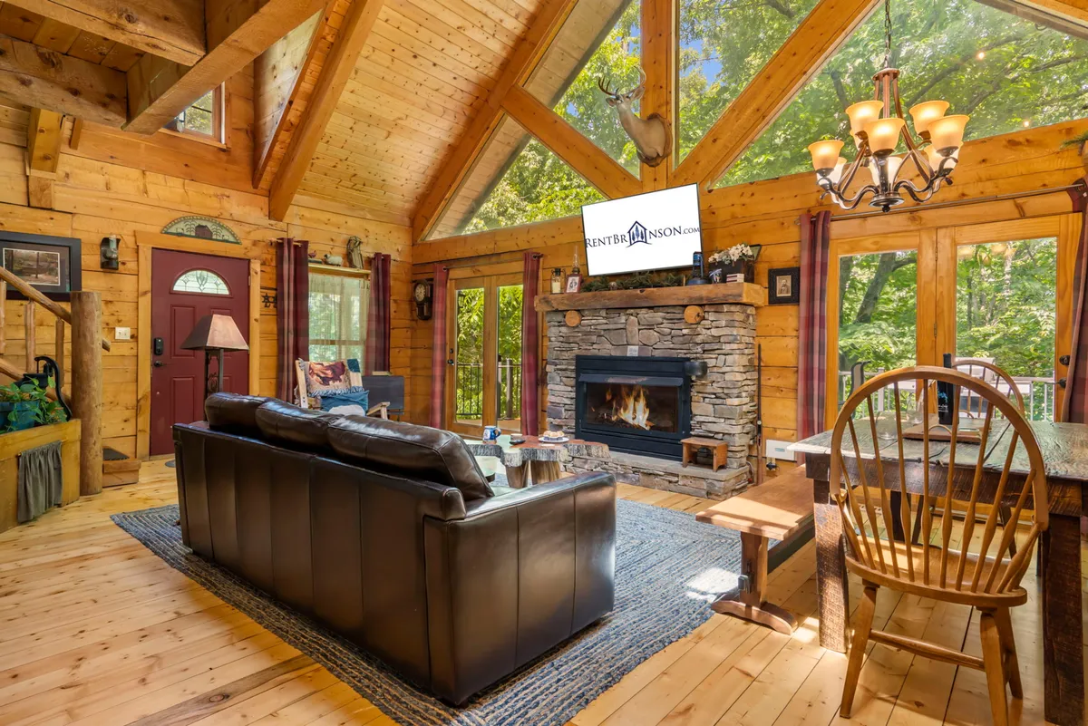 This image captures a spacious, light-filled living area featuring a striking stone fireplace, comfortable sofa, and large windows that overlook the outdoors. The dining area is visible, fostering a sense of openness and communal gathering.
