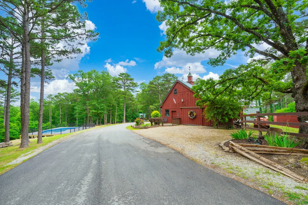 A peaceful exterior view featuring a classic red barn, lush trees, a gravel driveway, and a sparkling community swimming pool. The setting invites families to swim, walk, and relax in a communal space that blends into nature.