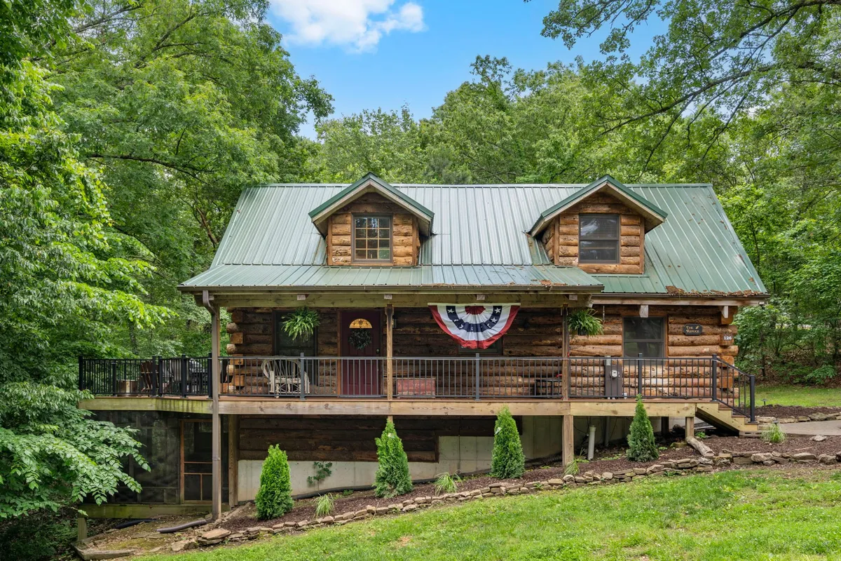 This image features the welcoming exterior of a log cabin with a green roof, a deck with railing, and festive decorative bunting. The surrounding natural landscape invites guests to enjoy the outdoors, with ample space for walks, play, and relaxation. The scene exudes a friendly, cozy atmosphere, ideal for families and their pets.