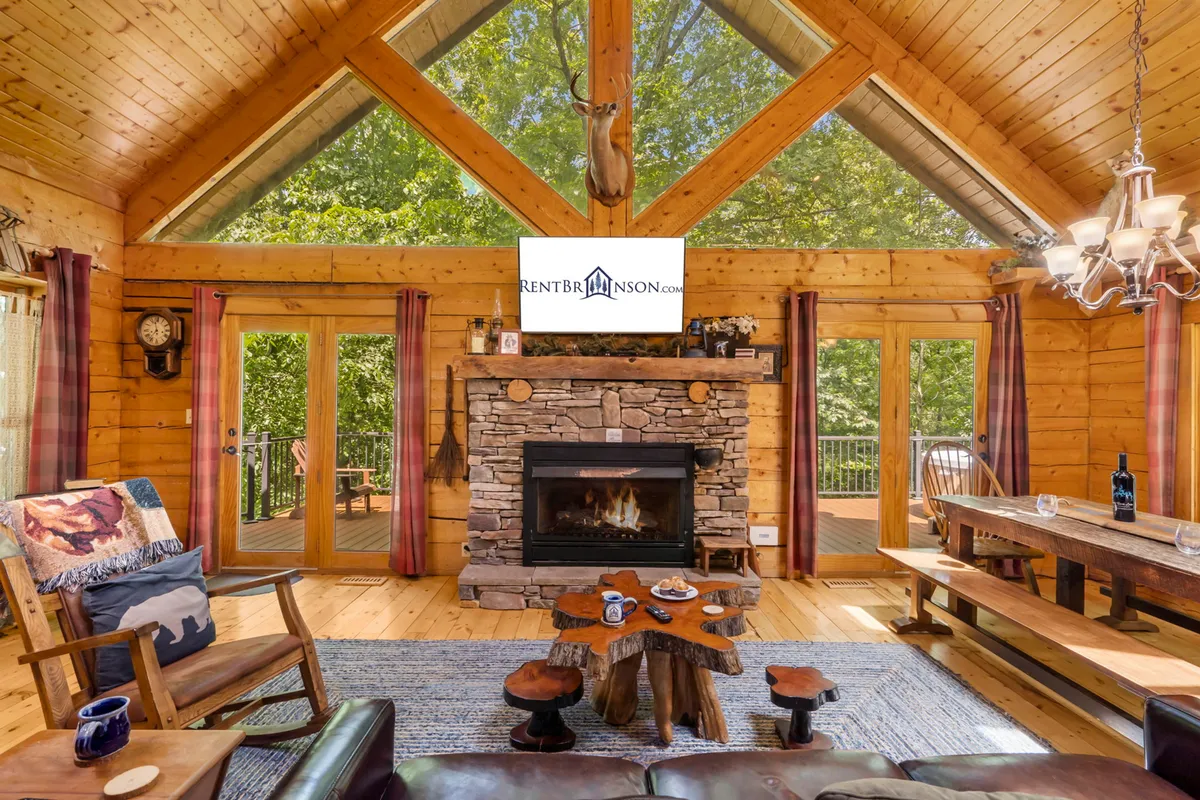 The image showcases the cozy interior of the Engleman Spruce Cabin, featuring a striking stone fireplace as the centerpiece, flanked by large windows that reveal wooded views. Warm wooden beams frame the open living space, while rustic furniture invites guests to relax and gather together. The setting exudes comfort and warmth, perfect for family nights by the fire or watching wildlife outside.