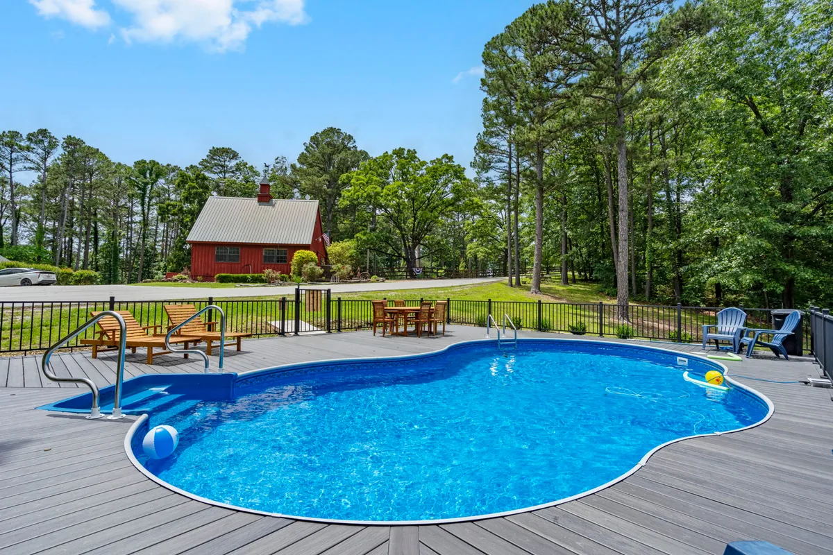 A sun-drenched swimming pool sits beside a red house, surrounded by a wooden deck and lounge chairs. The setup is perfect for group gatherings, pool parties, and relaxed sunbathing. The deck area offers ample space for friends or couples to enjoy drinks, laughter, and quality time outdoors.