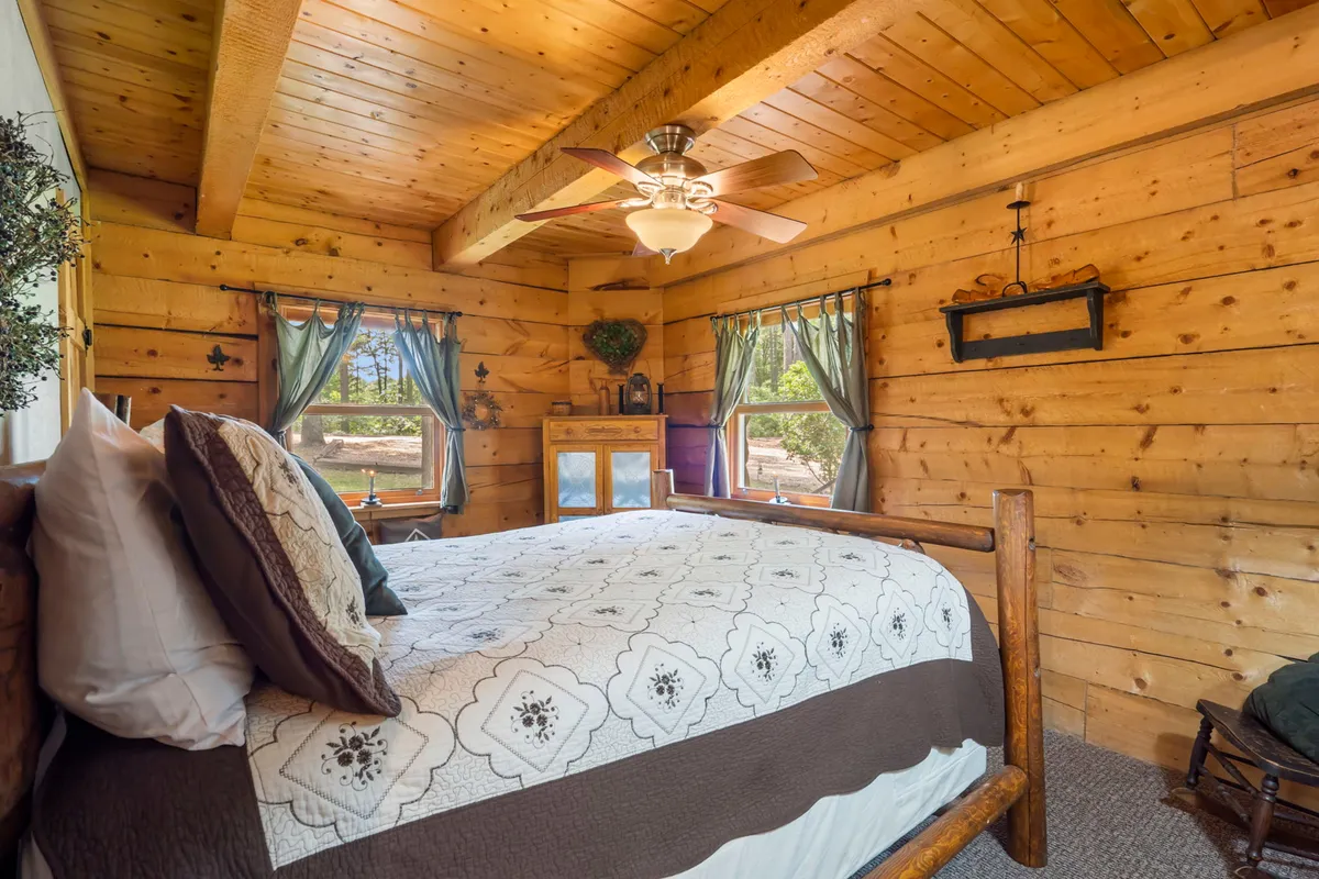 An inviting bedroom with a wooden bed, quilted bedspread, curtained windows, wooden ceiling, and ceiling fan. The room exudes warmth and coziness, with sunlight streaming in, perfect for curling up with a book or enjoying a peaceful sleep.