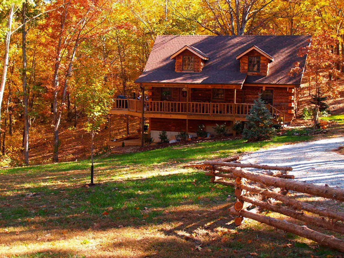 A cozy log cabin sits in front of vibrant fall foliage and a wooden fence, inviting guests with its front porch setting. The scene evokes tranquility and a strong connection to nature, emphasizing the peaceful and inviting atmosphere of the estate.
