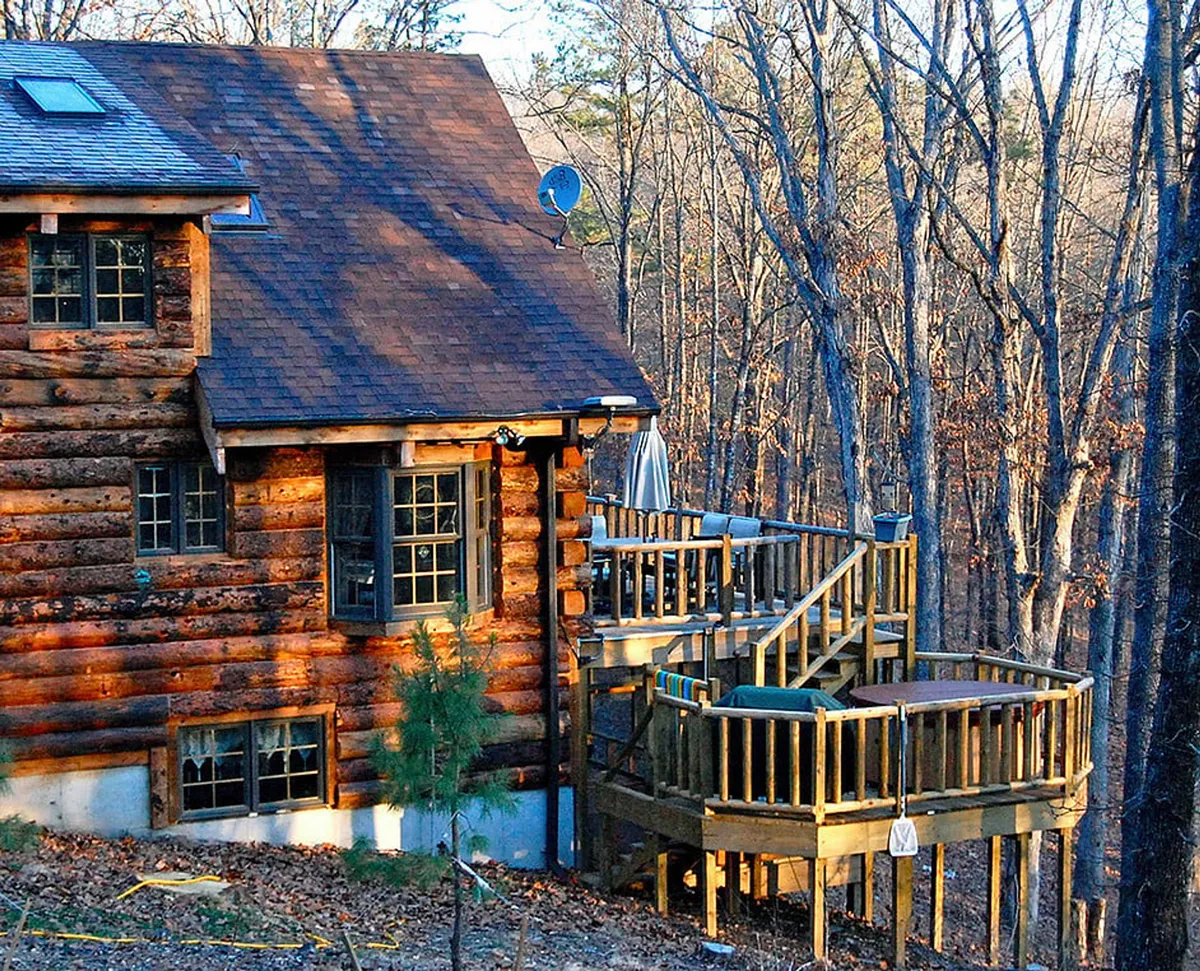 The image shows a tranquil log cabin structure with a wooden deck nestled among surrounding trees. The setting feels rustic and peaceful, with the deck providing a vantage point for wildlife spotting, relaxing, or sharing time outdoors with pets.