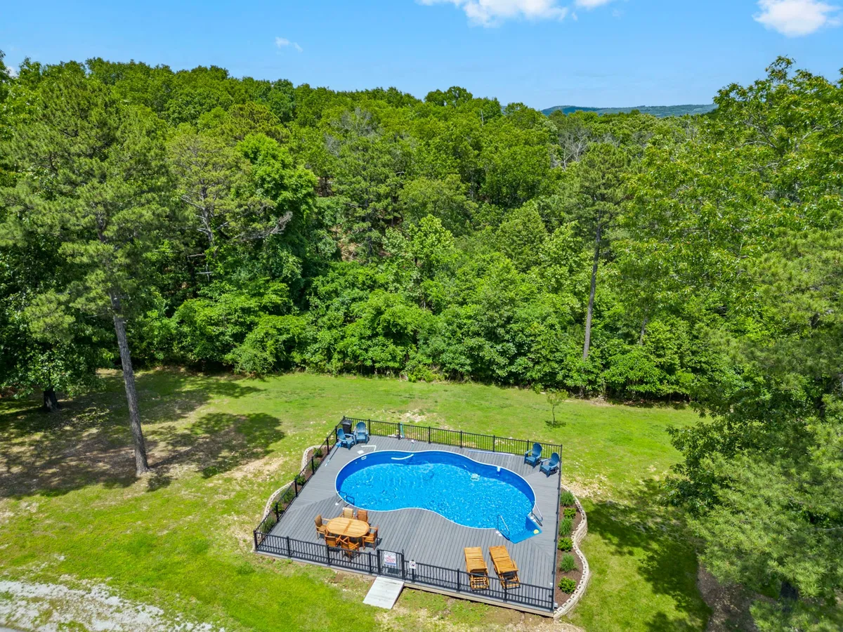 A bright exterior scene with a large swimming pool, deck area, lounge chairs, and surrounding trees. The setting is tranquil and well-maintained, ideal for sunbathing, swimming, and outdoor gatherings, all with a backdrop of lush Ozark greenery.