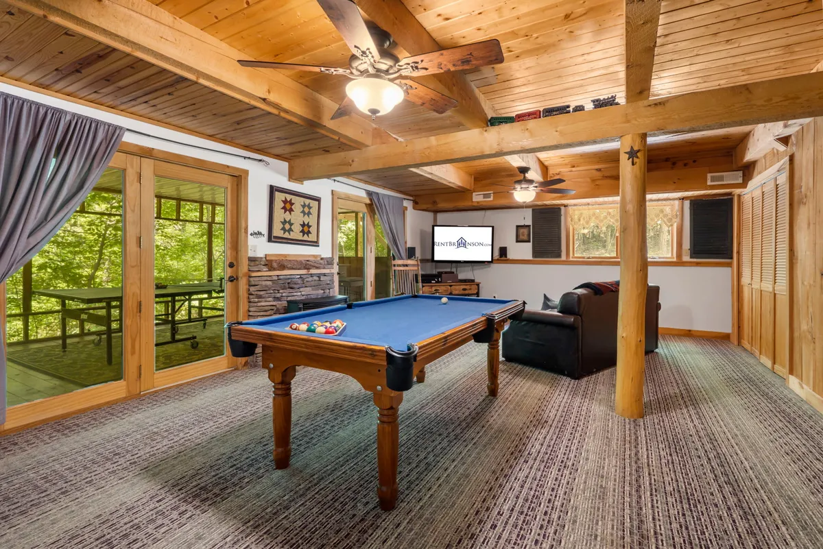 A spacious family room featuring a pool table as the focal point, complemented by a comfortable sofa, sliding glass doors letting in natural light, a television, and rustic wooden beams overhead. The room is styled for casual, social gatherings and leisure activities.