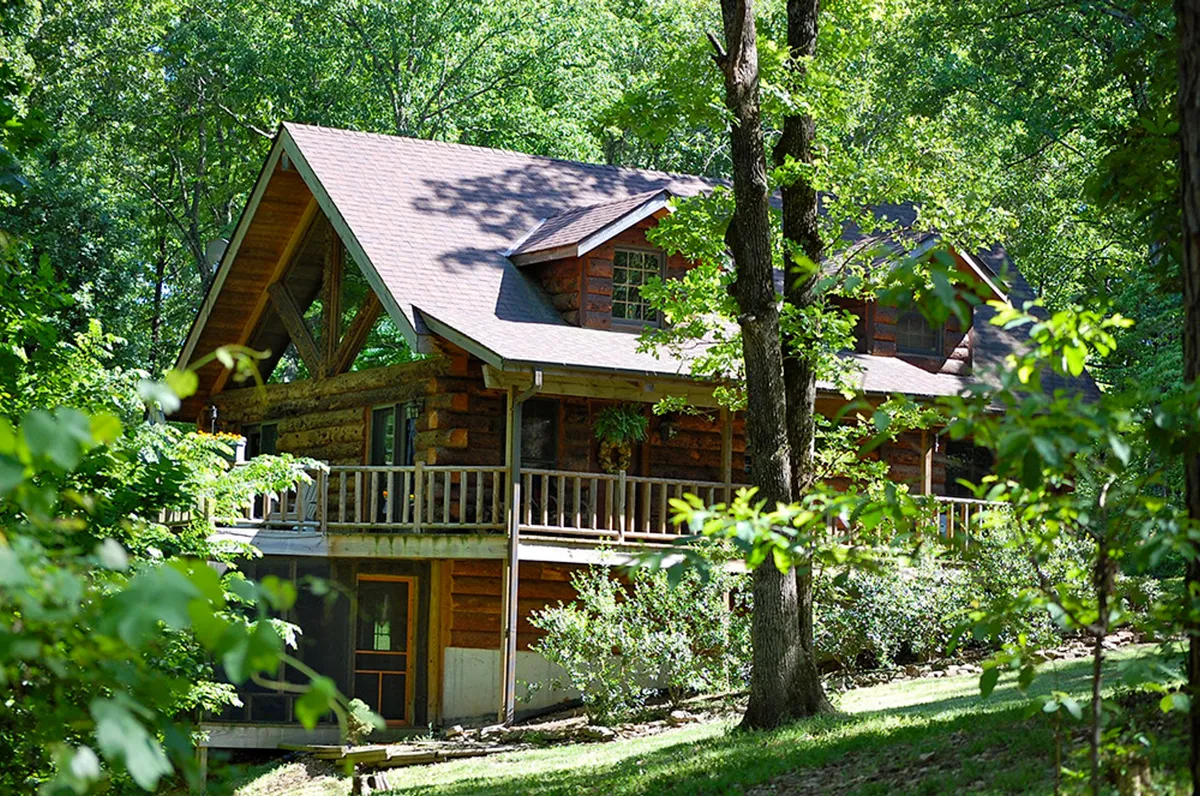 A log cabin with a large front porch nestled among tall, mature trees. The cabin is surrounded by lush greenery and well-kept landscaping, conveying a deep sense of seclusion and immersion in nature. The spacious porch hints at opportunities for outdoor relaxation, sunset watching, and stargazing. There is ample space for gatherings around a firepit, and the setting evokes tranquility and connection to the outdoors.