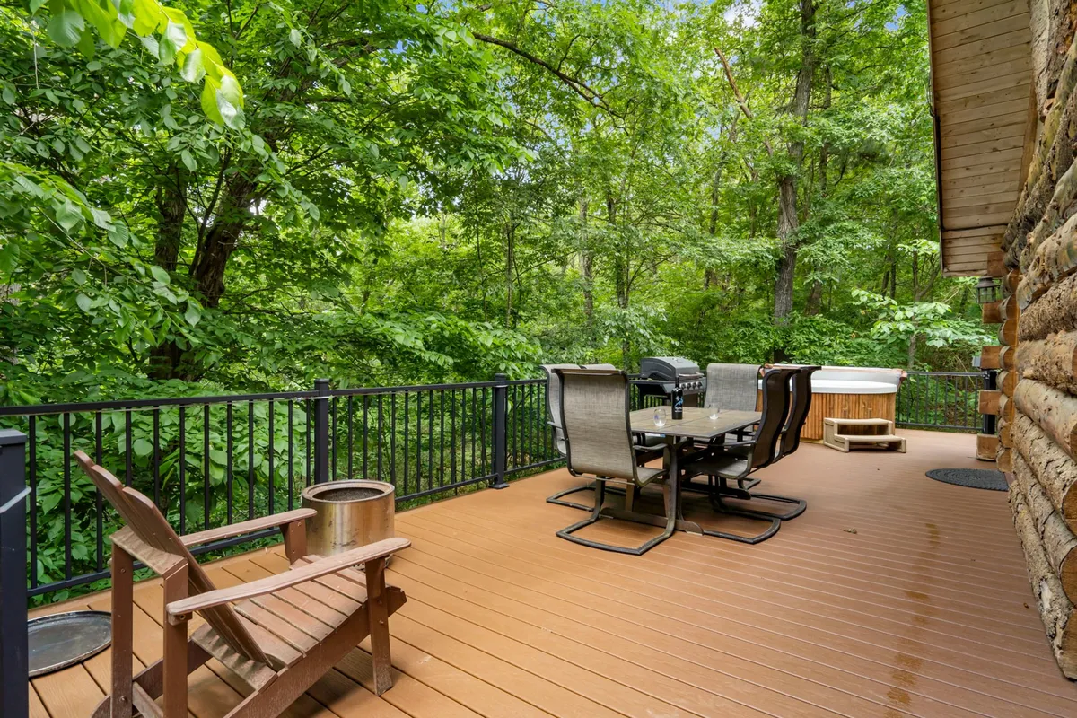 A spacious wooden deck with an inviting outdoor dining set, hot tub, and lush greenery surrounding the area. The scene exudes serenity, with the deck perfectly positioned for sunset views and stargazing, while the hot tub and seating provide options for both relaxation and social gatherings.