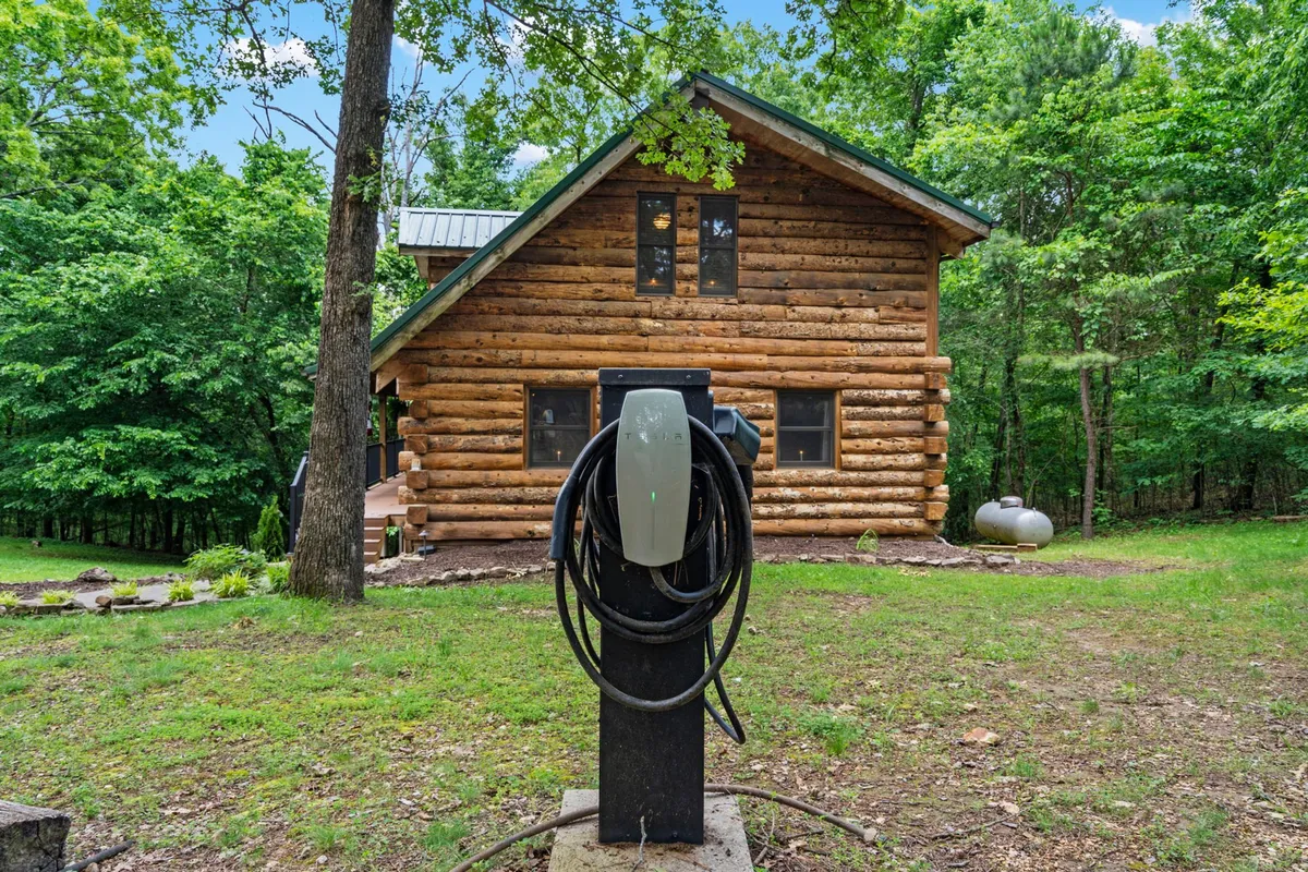 This exterior image highlights a log cabin nestled among trees, with a prominent electric vehicle charger visible next to the cabin. The natural setting showcases the peaceful surroundings and eco-friendly amenities offered on the estate.