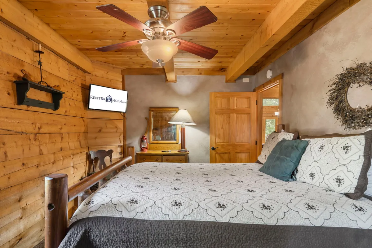 A cozy bedroom featuring a bed with a patterned quilt, a wooden ceiling, ceiling fan, wall-mounted TV, and a decorative wreath. The room is bathed in natural light, creating a peaceful and inviting atmosphere ideal for rest and relaxation.